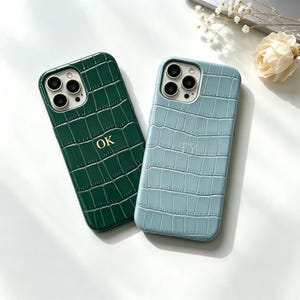Può includere: Due cover per smartphone con una texture a motivo pelle di coccodrillo. Una cover è verde scuro con le lettere dorate OK, l'altra è azzurra con le lettere dorate TY. Entrambe le cover presentano un'apertura per il sistema a tripla fotocamera.