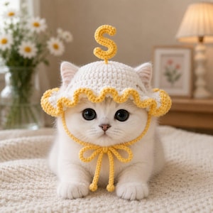 Pode incluir: Um gatinho branco usando um chapéu de crochê com borda amarela franzida e uma letra S amarela no topo. O chapéu possui uma tira amarela amarrada em um laço sob o queixo. O gatinho tem pelo branco e grandes olhos escuros, olhando para frente.