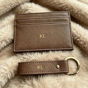 May include: A brown card holder and a matching rectangular keychain, both featuring a textured finish and the gold-embossed initials KL. The card holder has multiple slots, while the keychain includes a gold-toned metal ring and a small stud.