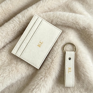 May include: A white rectangular card holder with multiple slots and a matching white keychain strap featuring a silver ring. Both items are made of a textured material and are embossed with the gold-colored capital letters KL on the front.