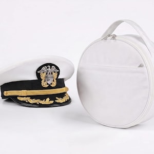 May include: A white and black military-style peaked cap featuring a gold chin strap and a silver eagle emblem, positioned next to a round, white, zippered carrying case with a top handle, both set against a plain white background.