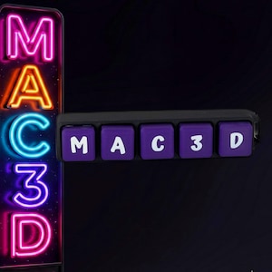 May include: A vertical neon sign displaying the text MAC3D in pink, orange, and blue light, positioned next to a black horizontal bar featuring five purple square keys with the white letters M, A, C, 3, and D printed on their surfaces.