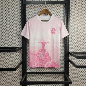 May include: A short-sleeved t-shirt in shades of light pink and white, featuring a graphic print of the Christ the Redeemer statue on Corcovado Mountain in Rio de Janeiro, with a small crest logo on the upper left chest area of the garment.