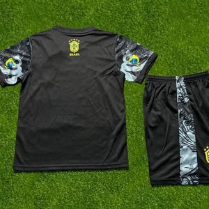 May include: A black short-sleeved athletic shirt and matching shorts set. The shirt features patterned sleeves with blue and yellow accents and a yellow Brasil crest with five stars. The shorts have a side panel with a graphic print and a yellow Brasil crest.