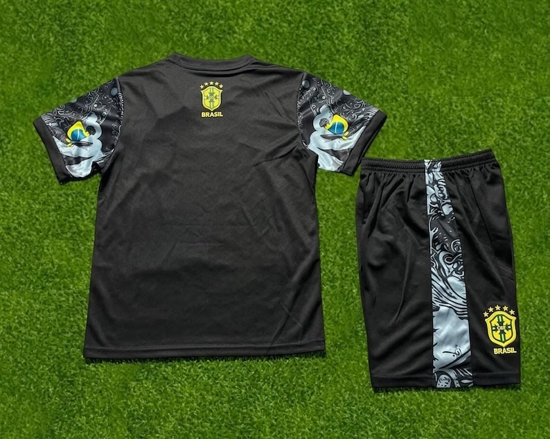 May include: A black short-sleeved athletic shirt and matching shorts set. The shirt features patterned sleeves with blue and yellow accents and a yellow Brasil crest with five stars. The shorts have a side panel with a graphic print and a yellow Brasil crest.