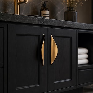 May include: Two matte gold, curved, crescent-shaped cabinet handles mounted vertically on black wooden cabinet doors. The handles feature a smooth, tapered design that contrasts with the dark, textured wood grain of the cabinetry.