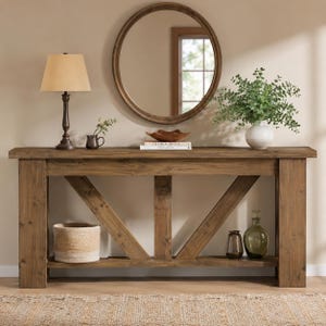 May include: A rectangular wooden console table with a natural brown finish, featuring a lower shelf and X-shaped support braces. The top surface holds a table lamp, a small pitcher, a wooden bowl, books, and a white vase with green foliage.