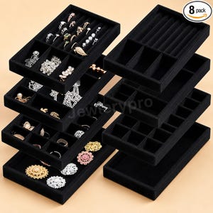 May include: A set of eight black velvet jewelry organizer trays with various compartment layouts, including ring slots and open sections. Some trays are filled with assorted rings, necklaces, and earrings, while others are empty.