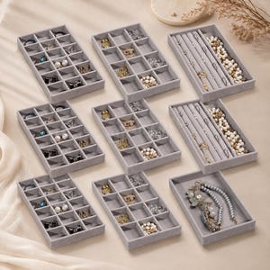 May include: A set of nine light gray velvet jewelry organizer trays arranged in three rows. Six trays feature multiple square compartments for small items, while three trays contain long, narrow slots for necklaces and bracelets.