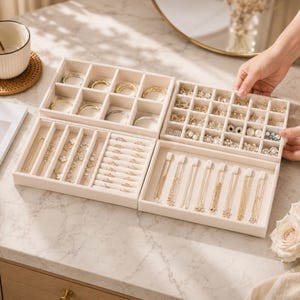 May include: A set of four modular beige velvet jewelry organizer trays. The trays feature various compartments for rings, earrings, necklaces, and bracelets. The organizers are filled with assorted gold and silver jewelry pieces, including chains, studs, and rings.