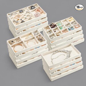 May include: A 16-pack of rectangular, off-white jewelry organizer trays with various internal compartment configurations. The trays are stacked in four groups and contain assorted rings, bracelets, necklaces, and earrings in gold and silver tones.