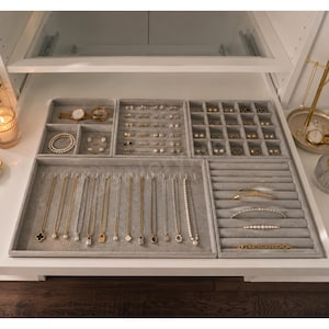 May include: A set of modular gray velvet jewelry organizer trays arranged inside a drawer. The trays feature various compartments for necklaces, rings, earrings, and bracelets, displaying gold and silver chains, pearl strands, and stud earrings.
