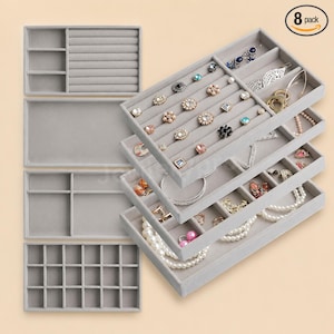 May include: An 8-pack of light gray velvet jewelry organizer trays featuring various compartment layouts, including ring slots, square grids, and open sections, displayed with assorted rings, necklaces, and earrings inside the trays.