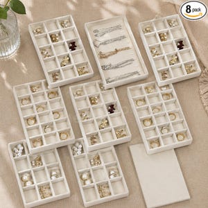 May include: A set of eight beige velvet jewelry organizer trays with multiple compartments for rings, earrings, and necklaces. The trays feature various gold and silver jewelry pieces, including chains, rings, and stud earrings, arranged within the grid sections.