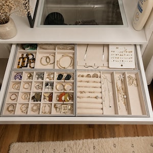 May include: A white jewelry drawer organizer with four velvet-lined trays containing various gold, silver, and tortoiseshell earrings, rings, necklaces, and bracelets. The compartments are filled with assorted jewelry pieces, including hoop earrings, stud earrings, and delicate chains.