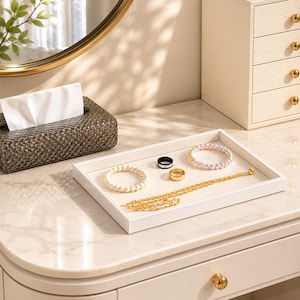 May include: A rectangular white jewelry tray holding a collection of accessories including a white beaded bracelet, a black ring, a gold-toned ring, a multi-colored pastel beaded bracelet, and a long gold-toned chain necklace with a small ball charm.