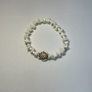May include: A stretch bracelet composed of irregular white stone chip beads and small round white beads. The centerpiece is a white turtle charm featuring a brown etched shell pattern. The beads are strung on an elastic cord to form a circular shape.