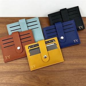 May include: Five rectangular card holders in black, blue, light blue, brown, and yellow. Each holder features a central vertical strap with a silver snap button, multiple horizontal card slots, and the gold embossed letters TY in the corner.