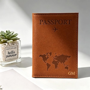 以下が含まれることがあります： 上部にPASSPORTの文字と小さな飛行機のアイコンが刻印された茶色のレザー製パスポートケース。中央には世界地図のグラフィックが刻印されており、右下隅にはGMのイニシャルが刻印されています。