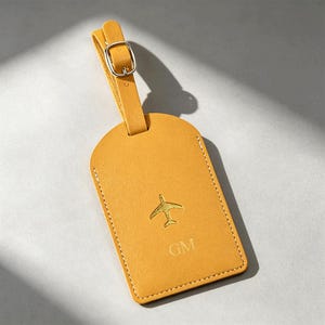 May include: A mustard yellow luggage tag with a gold-toned metal buckle strap. The tag features an embossed airplane icon centered above the letters GM, with visible white stitching along the rounded rectangular edges of the main body.