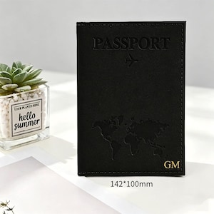 Op de afbeelding: Een zwarte paspoorthouder van 142 bij 100 mm. De voorzijde is voorzien van het in reliëf aangebrachte woord PASSPORT, een vliegtuigicoon, een wereldkaartsilhouet en de goudkleurige initialen GM. De hoes heeft zichtbare stiksels langs de randen.