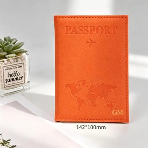 May include: An orange passport holder measuring 142 by 100 millimeters. The cover features the embossed word PASSPORT, an airplane icon, a world map graphic, and the gold-colored initials GM in the bottom right corner with visible stitching.