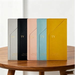 Può includere: Cinque portafogli da viaggio rettangolari in grigio, nero, rosa chiaro, azzurro e giallo senape. Ogni portafoglio presenta una tasca frontale diagonale, le lettere TY impresse in oro e il testo Travel Wallet Have a Nice Trip in basso.