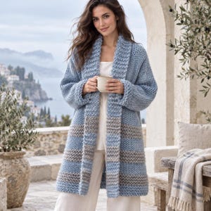 Puede incluir: Un cárdigan de punto grueso con cuello de chal y mangas largas. La prenda está diseñada con rayas horizontales en tonos alternos de azul claro y gris suave. Se lleva sobre una parte superior blanca lisa y pantalones blancos.