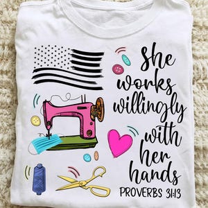 Könnte beinhalten: Ein weißes T-Shirt mit einem Grafikdruck einer rosa Nähmaschine, einer blauen Gesichtsmaske, einer gelben Schere, bunten Knöpfen, einer Garnrolle, einem rosa Herz und einer stilisierten amerikanischen Flagge. Der Text lautet: She works willingly with her hands PROVERBS 31:13.