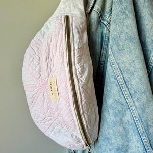 May include: A crescent-shaped quilted fanny pack in soft pink and white fabric with a beige strap. The bag features a vertical gold-tone zipper closure and a small rectangular fabric label that reads LINDSEY LOO DESIGN CO. in black text.