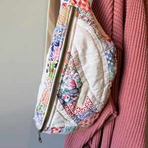May include: A quilted crossbody bag featuring a patchwork design of various floral and gingham fabric patterns in red, blue, and white. The bag has a long beige strap, a metal zipper closure, and a small white label reading LINDSEY LOO DESIGN CO.