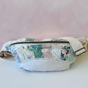 May include: A floral patchwork fanny pack with a cream-colored strap and zipper. The bag features a variety of floral patterns in shades of pink, blue, and green. The bag is made of quilted fabric.