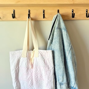 May include: A light pink quilted tote bag with cream-colored fabric handles and a matching ribbon tie, hanging alongside a light blue denim jacket on a wooden wall-mounted rack with black metal hooks, positioned below framed photographs.