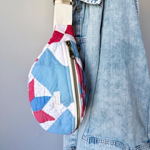 May include: A quilted fanny pack with a cream-colored strap and a bronze zipper. The bag features a patchwork design in shades of blue, red, and white. A denim jacket hangs in the background.