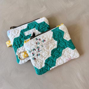 May include: Two quilted zipper pouches with white and teal geometric patterns. One pouch features a floral design with red berries. Each pouch has a black zipper and a small tag that reads "LINDSEY LOOP DESIGN CO."