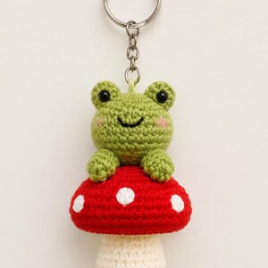 Peut inclure: Un porte-clés en crochet représentant une petite grenouille verte aux yeux noirs et aux joues roses, assise sur un champignon rouge à pois blancs. La grenouille est fixée à un anneau en métal argenté par une courte chaîne texturée.