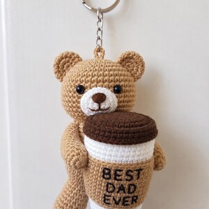 Op de afbeelding: Een gehaakte lichtbruine teddybeer sleutelhanger die een wit en beige koffiebeker met een donkerbruine deksel vasthoudt. Op de beker staat de zwarte geborduurde tekst BEST DAD EVER. De beer heeft zwarte kraaloogjes en een zilveren metalen ring.