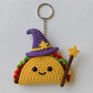 May include: A crochet taco keychain featuring a yellow shell, green lettuce, and red filling. The taco wears a purple pointed hat with a yellow star and holds a brown wand topped with a yellow star. It has two black bead eyes and a stitched smile.