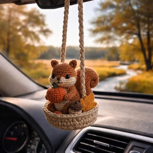 Peut inclure: Un ornement en crochet représentant un écureuil suspendu par une cordelette beige tressée. L'écureuil marron est assis dans un petit panier beige et tient une citrouille orange miniature, un gland marron et une feuille d'érable jaune.