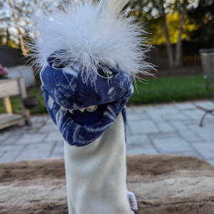 May include: A blue and white puppet with a white furry head and a long white arm. The puppet has a toothy grin and large black eyes.