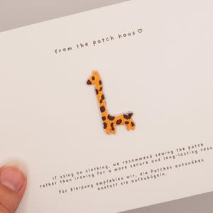 May include: A small giraffe-shaped embroidered patch with yellow and brown spots attached to a white card. The card features the text from the patch haus and instructions in English and German regarding sewing the patch onto clothing.
