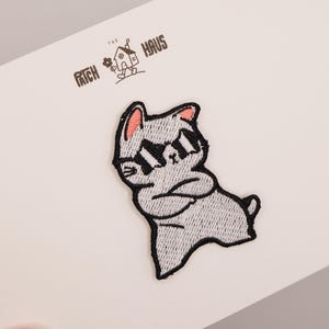 Puede incluir: Un parche bordado que muestra un gato gris con el interior de las orejas rosa y gafas de sol negras, de pie con los brazos cruzados. El parche está sujeto a una tarjeta blanca impresa con el texto THE PATCH HAUS y un icono de casa.