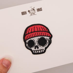 Könnte beinhalten: Ein gestickter Aufnäher mit einem weißen Totenkopf, großen schwarzen Augenhöhlen und einer roten Mütze. Der Aufnäher ist auf einer weißen Karte befestigt, auf der oben mittig der Text THE PATCH HAUS und eine kleine Hausgrafik schwarz gedruckt sind.