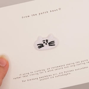 Op de afbeelding: Een witte geborduurde patch in de vorm van een kattengezicht met zwarte stiksels voor de ogen, snorharen en neus. De patch is bevestigd op een witte kaart met de tekst from the patch haus en instructies voor het opnaaien van de patch.