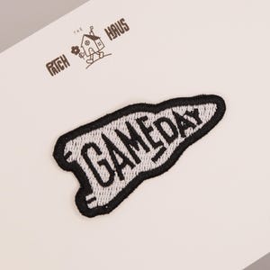 Op de afbeelding: Een wimpelvormige geborduurde patch met het woord GAMEDAY in zwarte draad op een witte achtergrond met een dikke zwarte rand. Boven de patch staat de tekst THE PATCH HAUS gedrukt met een kleine illustratie van een huisje dat een bloem vasthoudt.