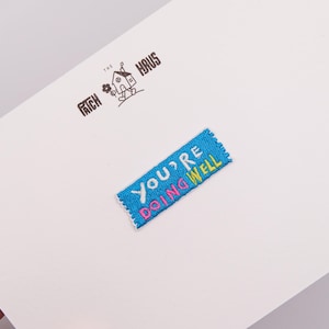 Può includere: Una toppa rettangolare blu ricamata con il testo "YOU'RE DOING WELL" in filo bianco, rosa e giallo. Sopra la toppa, un logo nero mostra una piccola casa con un fiore e il testo "THE PATCH HAUS" su un cartoncino bianco.