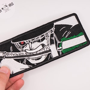 Pode incluir: Um emblema bordado retangular com uma ilustração gráfica em preto e branco de uma personagem segurando uma espada na boca. O design inclui um detalhe de olho vermelho e uma seção texturizada verde, tudo contornado por uma borda preta espessa.