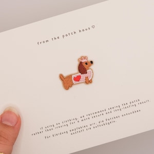 Pode incluir: Um emblema bordado de um cão salsicha castanho com uma coroa cor-de-rosa e uma camisola branca com um coração vermelho. O emblema está fixado num cartão branco com o texto from the patch haus e instruções para coser na roupa.