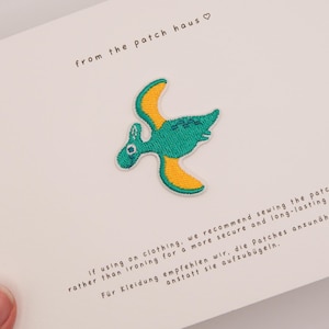 May include: An embroidered pterodactyl patch featuring a green body and yellow wings, centered on a white card. The card displays the text from the patch haus and instructions recommending sewing the patch onto clothing for a secure result.
