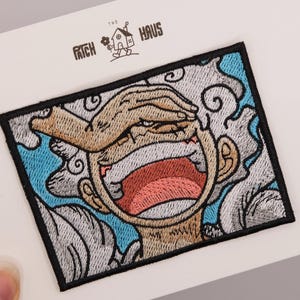 May include: A rectangular embroidered patch featuring a stylised character with a wide-open mouth, one hand covering their eyes, and swirling light blue and grey cloud-like patterns in the background. The patch has a thick black border.